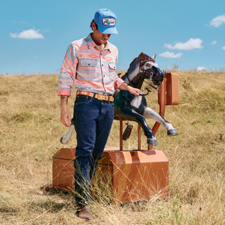 Man in a plaid shirt and blue cap standing next to a wooden horse statue in a grassy field.