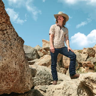 Man in a cowboy hat and patterned shirt standing on rocky terrain with a blue sky.