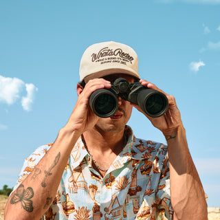 Person wearing a cap and patterned shirt using binoculars against a blue sky.