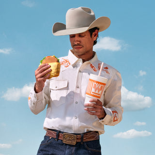 Man in cowboy hat holding a Whataburger and a drink against a blue sky with clouds