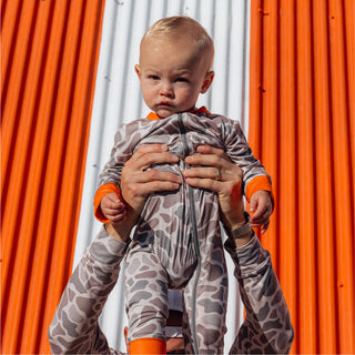 Baby in a camo Whataburger onesie being held up in front of an orange and white corrugated wall.