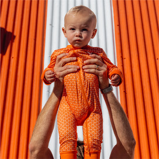 Baby in an orange Whataburger-patterned onesie being held up in front of an orange and white striped wall.