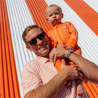 Smiling man in matching orange Whataburger polo holding a baby in coordinating orange onesie, both in front of a striped wall.