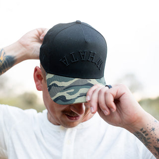 Close-up of a man adjusting his black Whataburger x True Brvnd cap with a camouflage-patterned brim. The brim partially shields his face, highlighting the sleek embroidered 'WHATA' embroidery on the front.