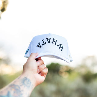 Hand holds up a white Whataburger x True Brvnd cap against a natural outdoor background. The cap features bold black embroidered lettering spelling an upside down, curved 'WHATA' across the front, with subtle branding details on the sides.