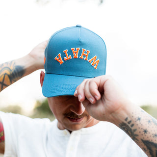 Close-up of a man adjusting blue Whataburger x True Brvnd cap. The brim partially shields his face, highlighting the orange and white embroidered 'WHATA' embroidery on the front.