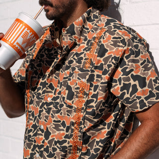 Person wearing a patterned shirt holding a cup with a straw against a white background