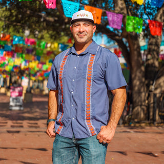 Man wearing a blue shirt with orange patterns and a white cap, standing in front of colorful paper flags.