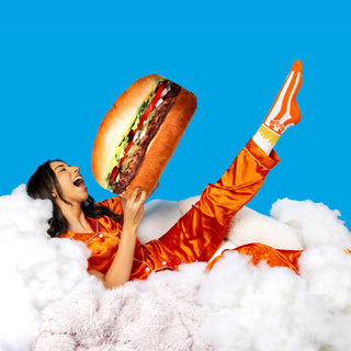 Smiling woman in orange satin pajamas lounges on fluffy clouds while playfully holding a giant Whataburger #1 Burger Pillow in front of a vibrant blue background.