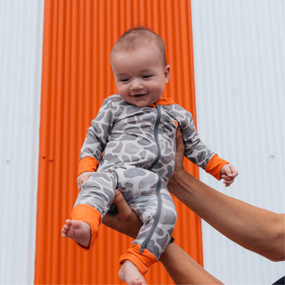 Baby held up in front of orange and white striped building wearing WHATA Classic Deer Camo Baby Zip Up.