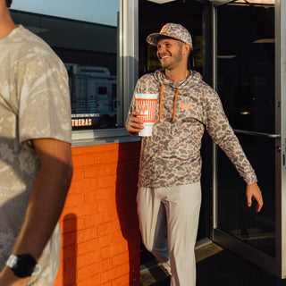 A man wearing a Classic Deer Camo performance hoodie with orange drawstrings and a matching cap walks out of a building while holding a Whataburger cup. Another person is partially visible in the foreground.