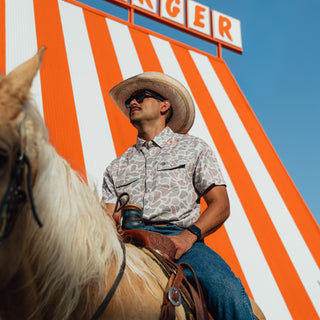 A man wearing a cowboy hat, sunglasses, and a patterned short-sleeve button-up shirt rides a light-colored horse in front of an orange and white striped Whataburger and a blue sky.