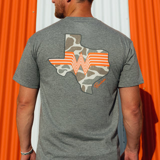 A man viewed from behind wearing a heather gray t-shirt with a graphic of the state of Texas filled with a camouflage pattern and orange stripes, set against a background of orange and white corrugated metal siding.