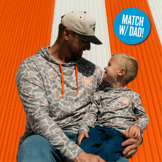 Child smiling while sitting on dad's lap wearing Youth WHATA Classic Deer Camo Performance hoodie in front of orange and white striped Whataburger a-frame building with circular callout reading "Match with Dad!"