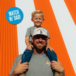 Child wearing Youth WHATA Texas Tee sitting on shoulders of dad wearing matching tee in front of orange and white striped Whataburger A-Frame building with circle sticker calling out "Match with Dad!"
