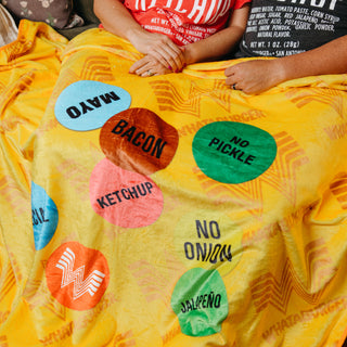 Yellow Whataburger burger-wrapper blanket with colorful topping labels draped across two people’s laps.