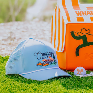 Light blue cap with 'Cowboy Golf' branding, orange bag with graphic design, and golf ball on grass.