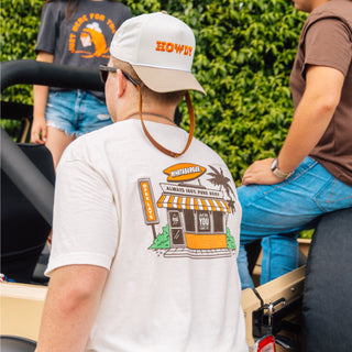 Person wearing a white t-shirt with a graphic design and a cap with text, sitting outdoors.