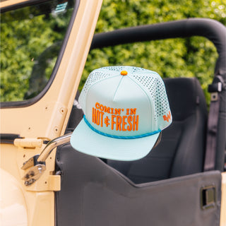 Light blue cap with orange text 'Comin' in Hot & Fresh' on the mirror of a vehicle.