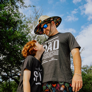 Father wearing Neon Collage Straw Hat, WhataDad Ingredient Tee, and Neon Collage swim trunks while looking down at son in front of trees and a blue sky.