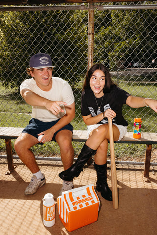 Two people sitting on a wooden bench at a baseball field, with a visible chain-link fence and trees in the background. An orange and white Whataburger cooler sits in the foreground, along with a drink bottle and a wooden bat.