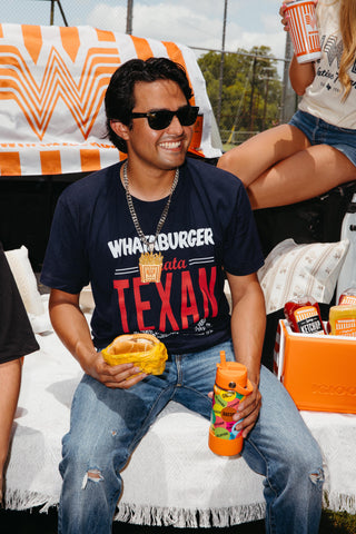 Man sitting on tailgate wearing Whataburger WhataTexan tee and Fry necklace, holding burger and water bottle. 