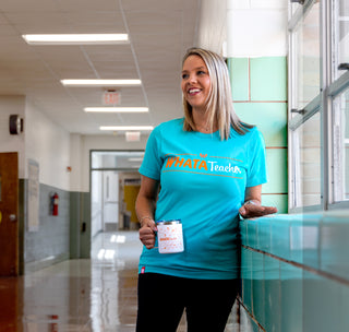 view woman wearing a sea green whatateacher tee holding a whatateacher mug standing in a school hallway.